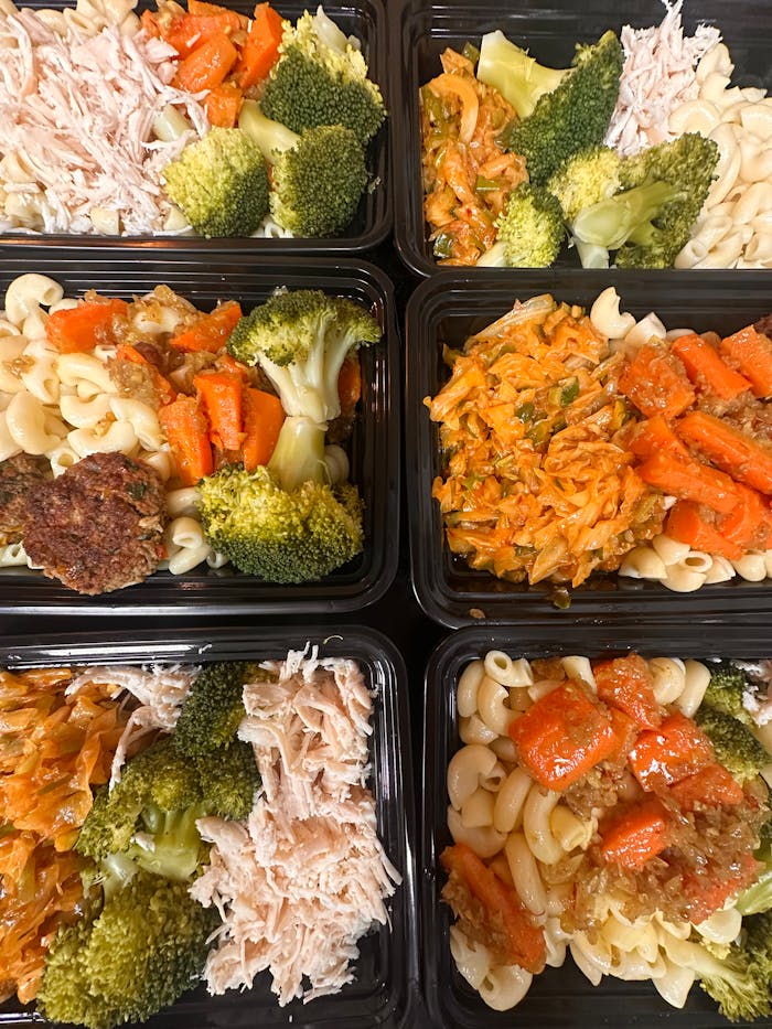 Colorful meal prep trays with pasta, broccoli, shredded chicken, and carrots, ready for healthy eating.